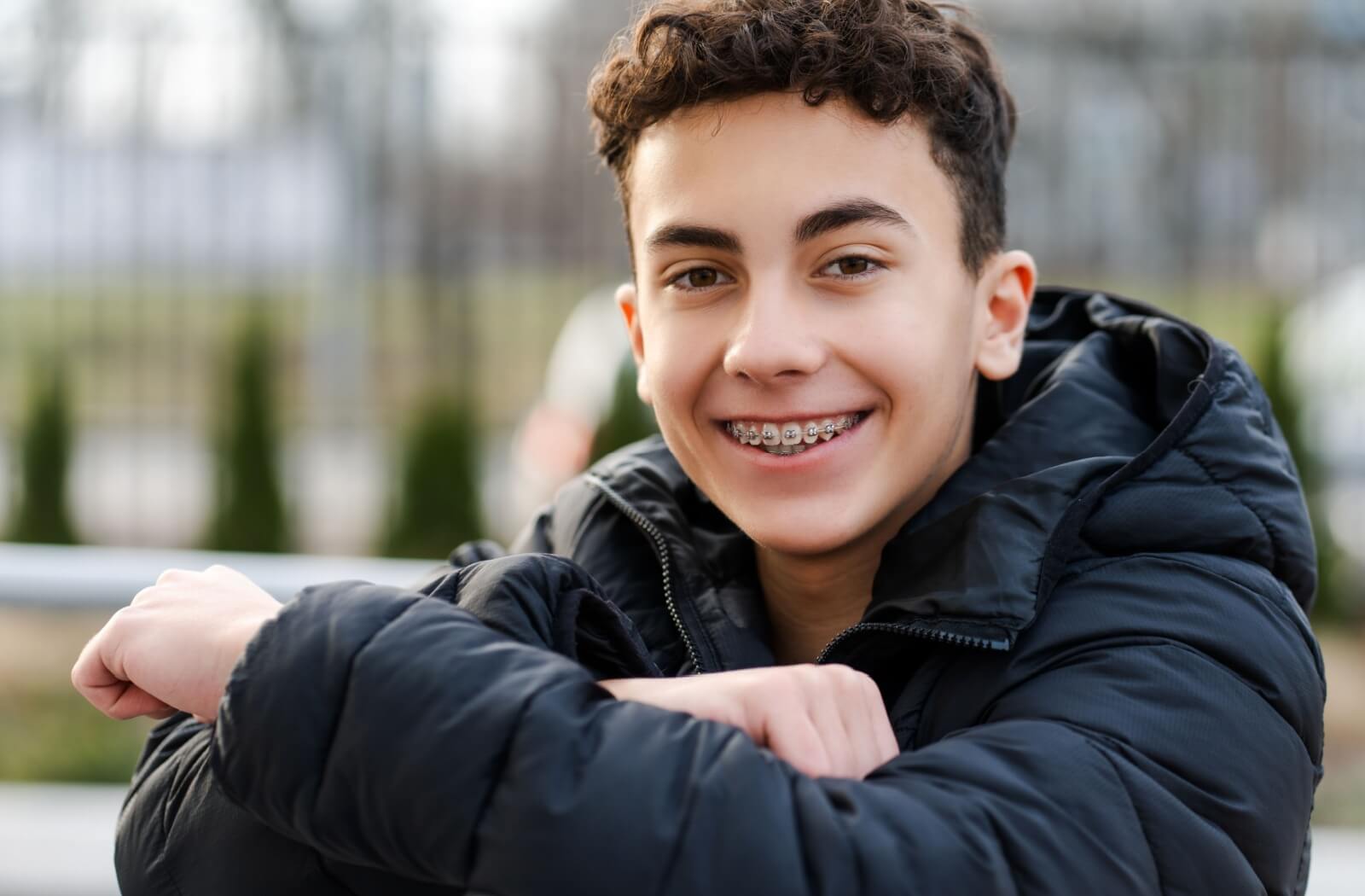 A smiling teenager wearing metal braces, sitting outside in a relaxed pose.