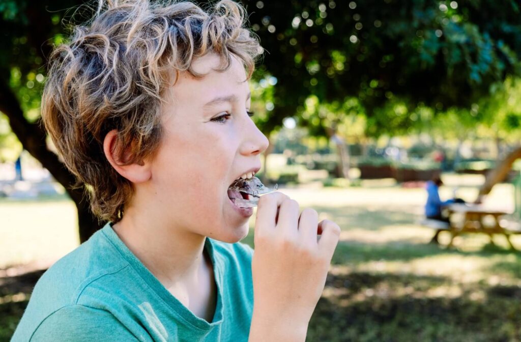 A young child outdoors, inserting removable aligners into their mouth.
