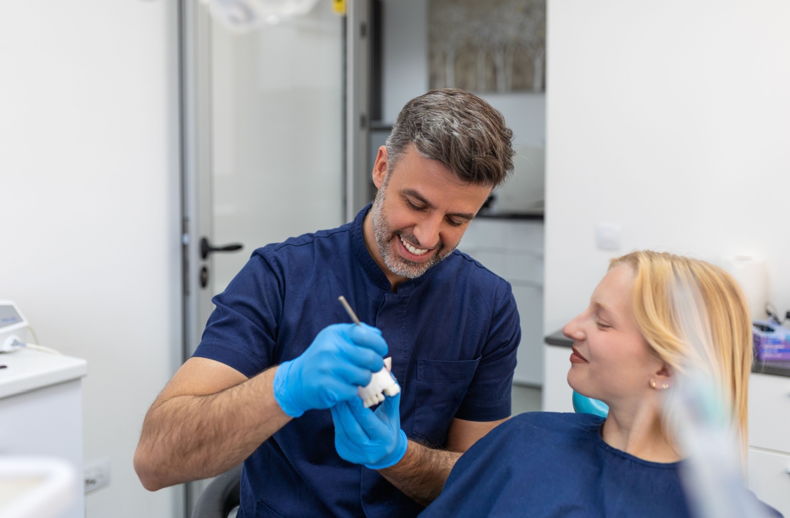 A dentist wearing gloves shows a model of a dental bridge to a patient in a dental chair.